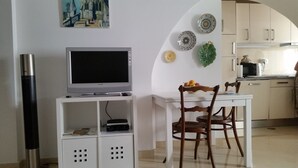 A dining area with a white table, two wooden chairs, a television on a white stand, and a kitchen in the background.