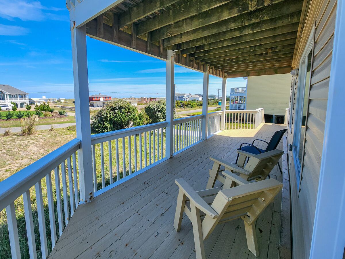 A covered porch with two wooden chairs and a table, overlooking a grassy area with distant buildings.