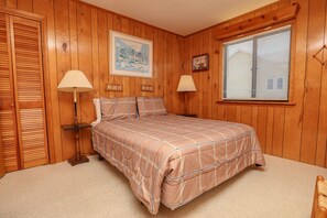 Ein Schlafzimmer mit Holzvertäfelung, einem Bett mit kariertem Überwurf, zwei Nachttischlampen, einem gerahmten Bild an der Wand und einem Fenster mit Jalousien.