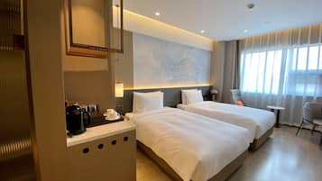 A hotel room with two beds, a desk with a coffee maker, a map on the wall, and a view of the city through the window.