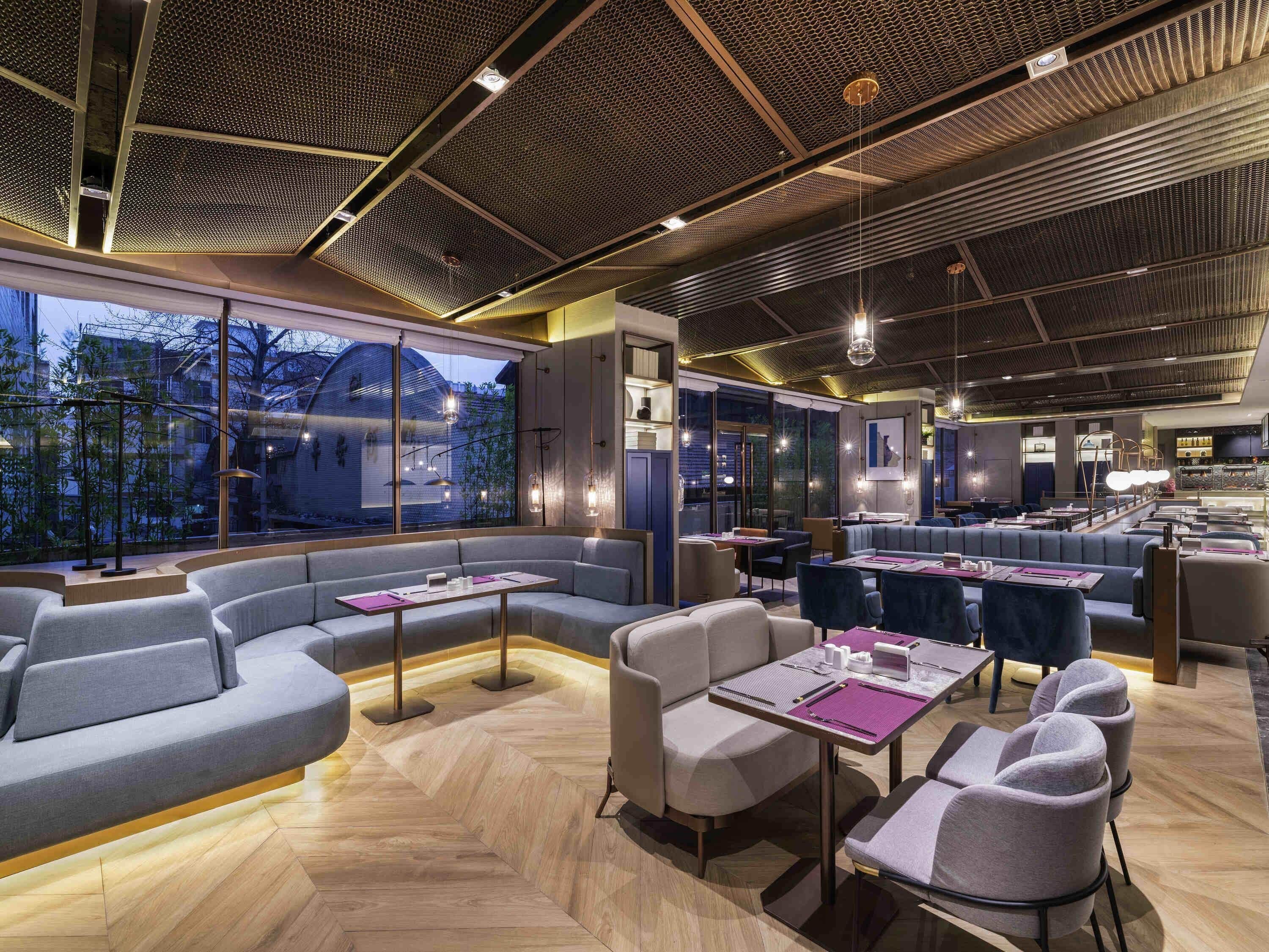 Een modern restaurant met een hoge plafond, grote ramen en diverse zithoeken, waaronder banken en tafels.