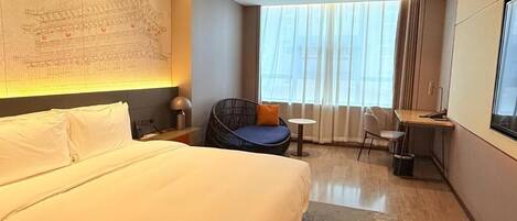 A hotel room with a large bed, a seating area with a chair and table, a desk, a large window with curtains, and a wall with a map.