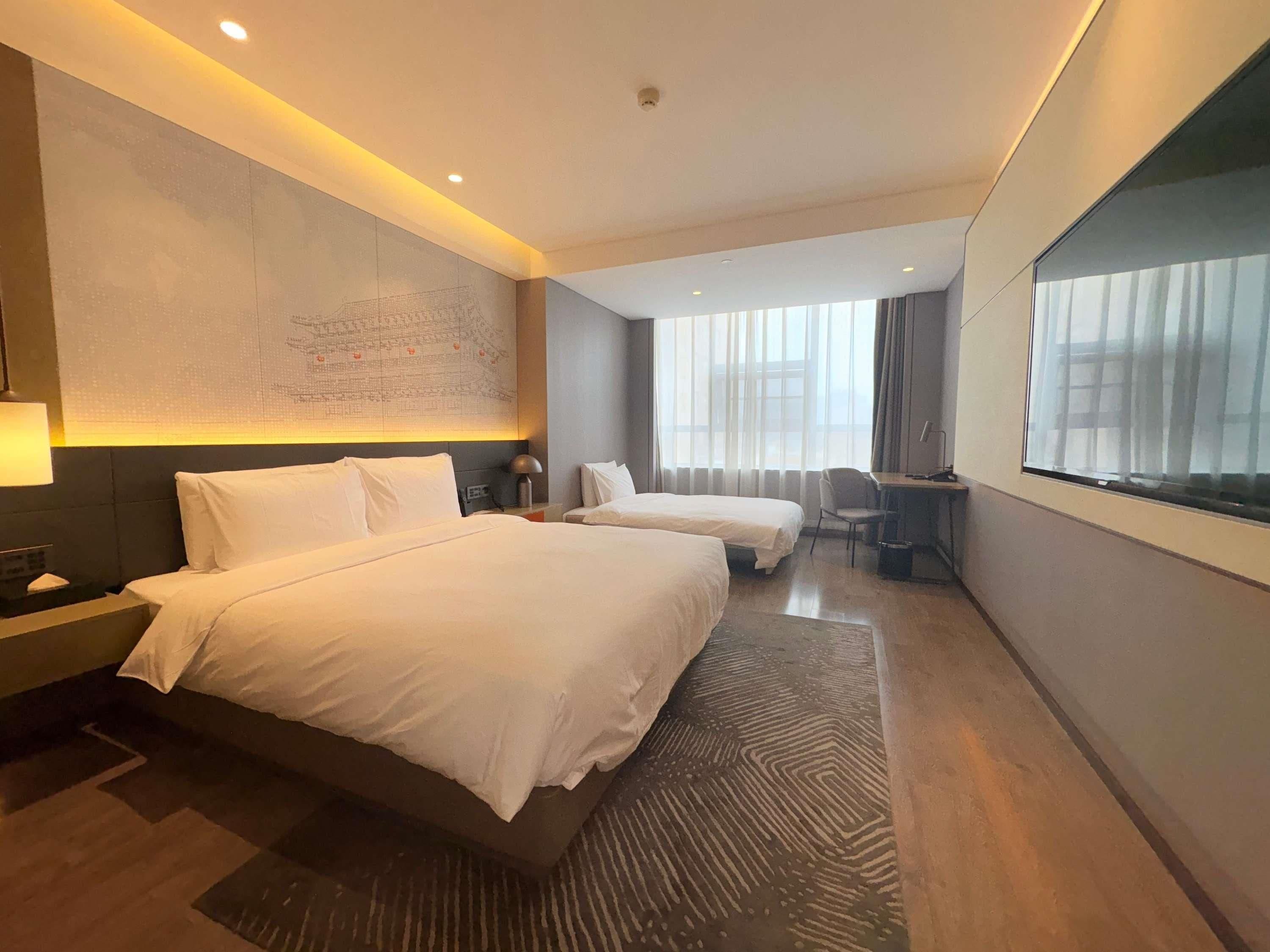 A hotel room with two beds, a desk, a chair, and a large window with curtains.