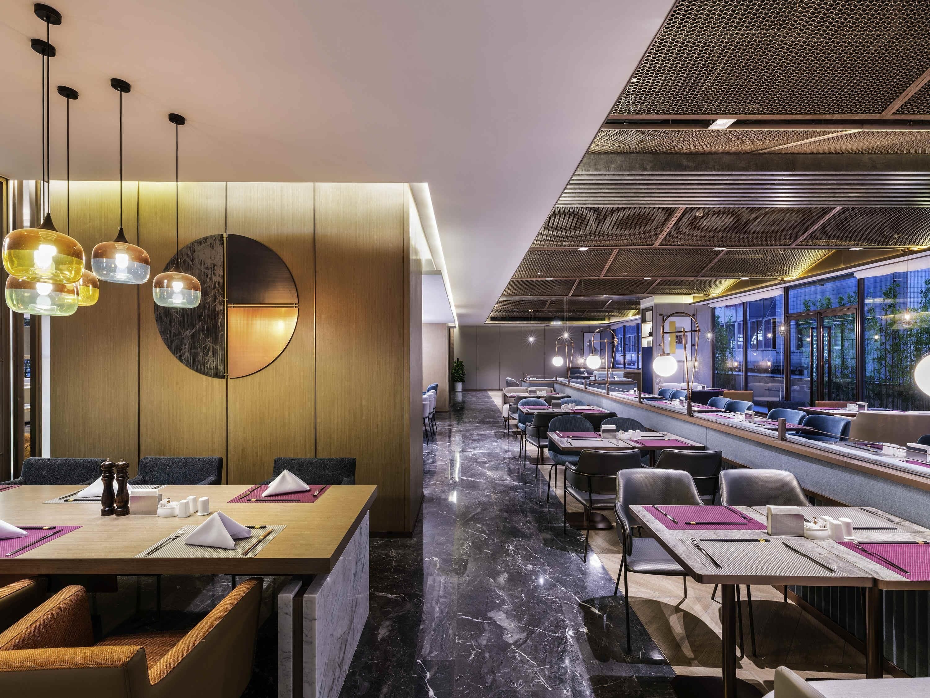 A modern restaurant with a long counter, multiple tables, and a high ceiling with a patterned design.
