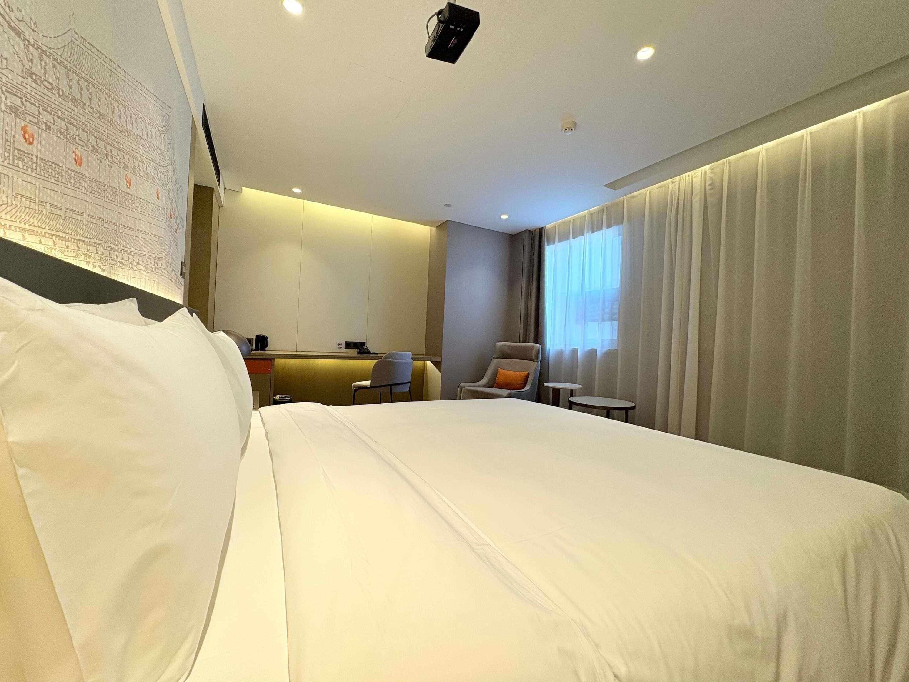 Habitación de hotel con una cama grande, un escritorio, dos sillas, una mesita pequeña y un ventanal con cortinas.