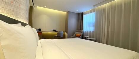 A hotel room with a large bed, a desk, two chairs, a small table, and a large window with curtains.