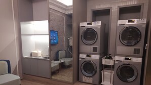 A laundry room with four stacked front-loading washing machines, a small seating area with a chair and table, and a television mounted on the wall. - Mercure Hangzhou East Railway Station (Hangzhou)