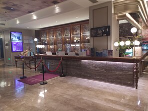 Una zona de recepción de hotel con mostrador de mármol, armarios de madera y una pantalla digital.