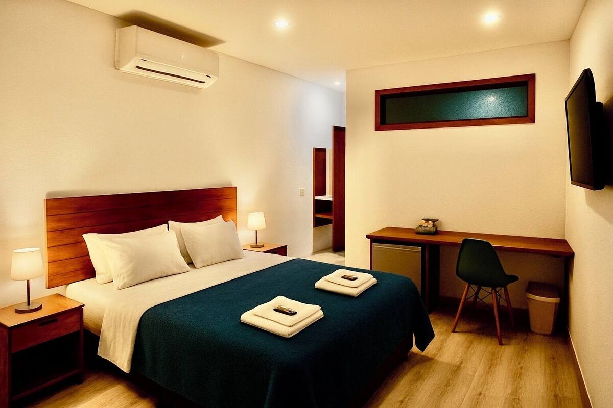 Una habitación de hotel con cama, mesitas de noche, escritorio, silla, televisión y aire acondicionado.