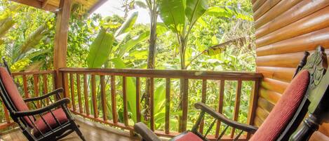 A wooden porch with two rocking chairs and a wooden railing, overlooking lush greenery.