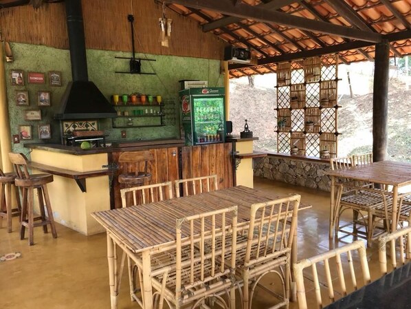 Uma área de bar ao ar livre rústica, com balcão de madeira, churrasqueira e frigorífico. Há mesas e cadeiras de vime, e uma parede de pedra ao fundo.