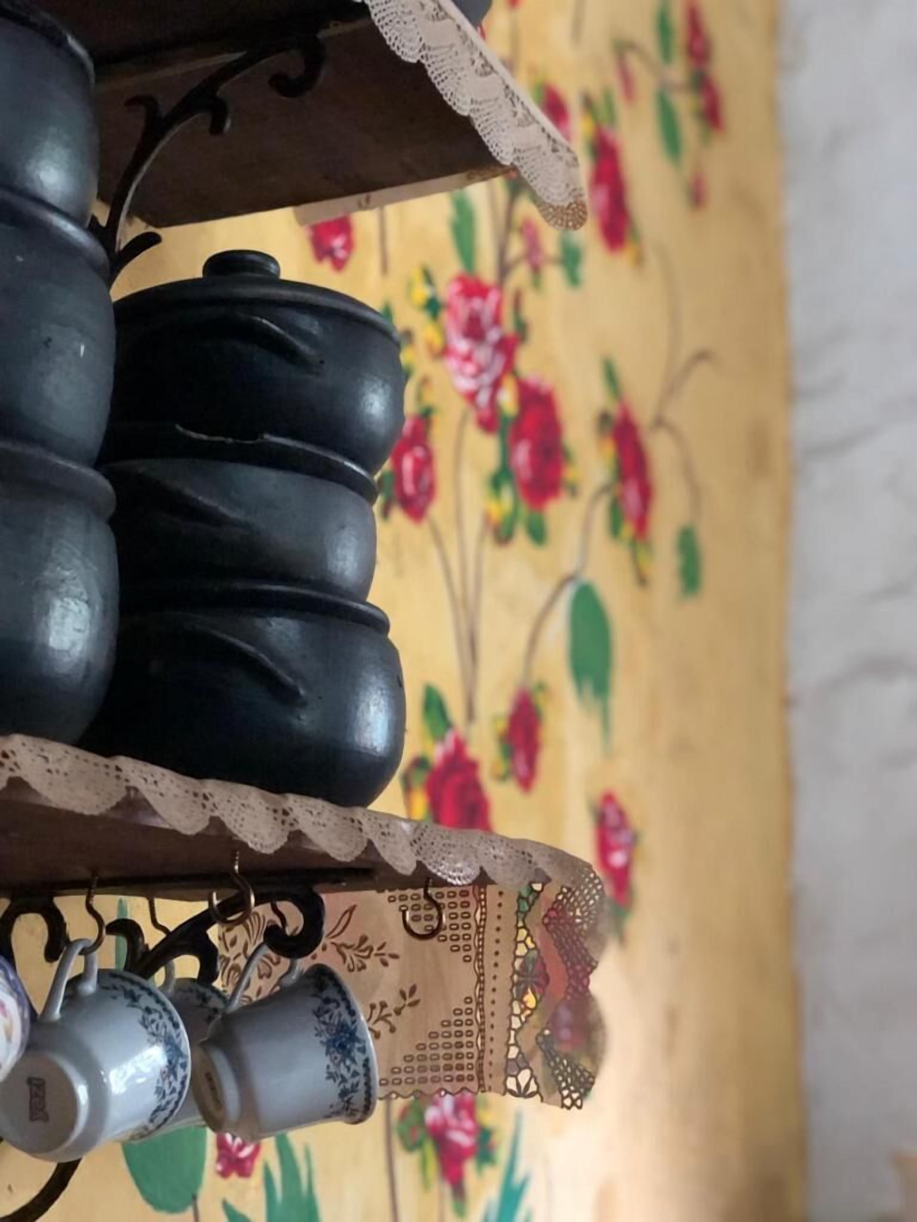 Primer plano de una maceta de cerámica negra sobre una estantería con encaje, sobre un fondo de papel tapiz floral.
