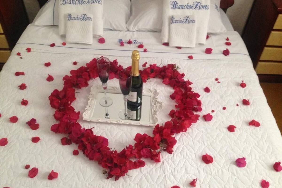 A bed with a heart-shaped arrangement of rose petals, a bottle of champagne, and glasses on a white quilted bedspread.