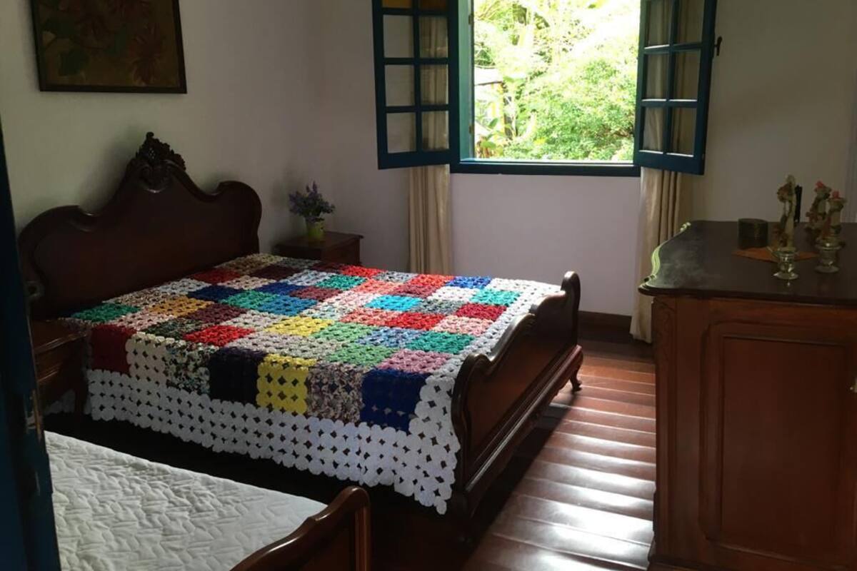 A bedroom with a wooden bed, a colorful quilt, a window with curtains, and a small table with a vase.