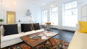 Un salon moderne avec un canapé blanc, une table basse sur laquelle sont posés des livres, un tapis coloré et de grandes fenêtres offrant une vue sur la ville.