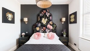Ein Schlafzimmer mit dunklem Kopfteil, Blumenbildern an der Wand, zwei Nachttischen mit Lampen und einem großen Fenster mit Jalousien.