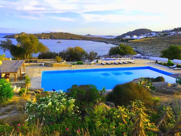 A pool with lounge chairs, a gazebo, and a view of the sea and hills.