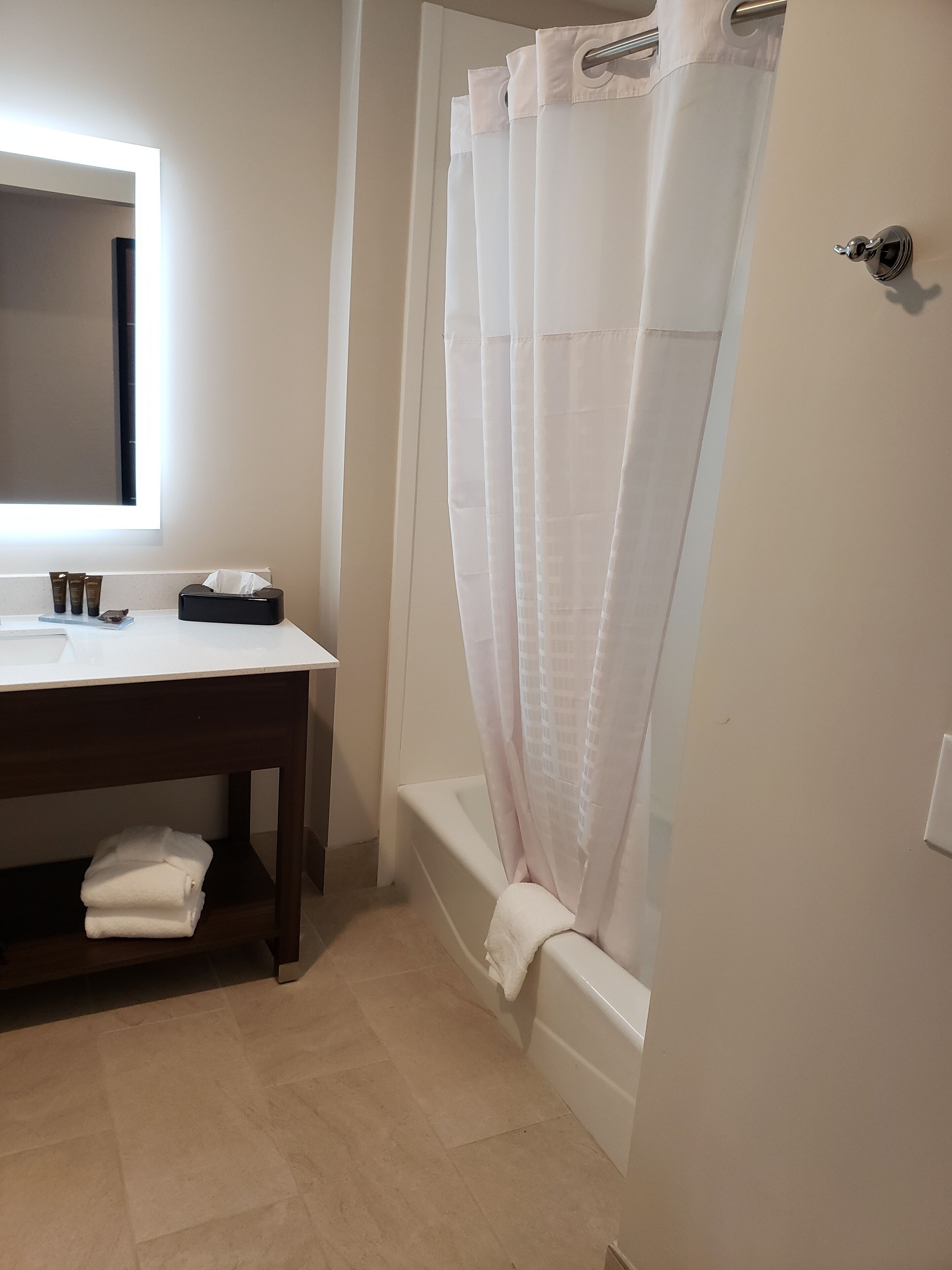 Une salle de bain avec un lavabo blanc, un miroir et une baignoire avec un rideau de douche. On y trouve un porte-serviettes et une petite étagère avec des articles de toilette.