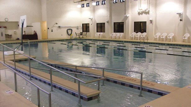 Una piscina interior con carriles delimitados, una silla de salvavidas y una zona junto a la piscina con sillas.
