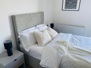 Cozy retreat with a grey upholstered headboard and white bedding, featuring a bedside table and framed sign with notable Parisian locations.