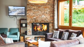 A cozy living room with a fireplace, a television mounted on the wall, and a large window with a view of greenery. - Chalet Mélèzor, sleeps 15, Serre Chevalier 1350 (Saint-Chaffrey)