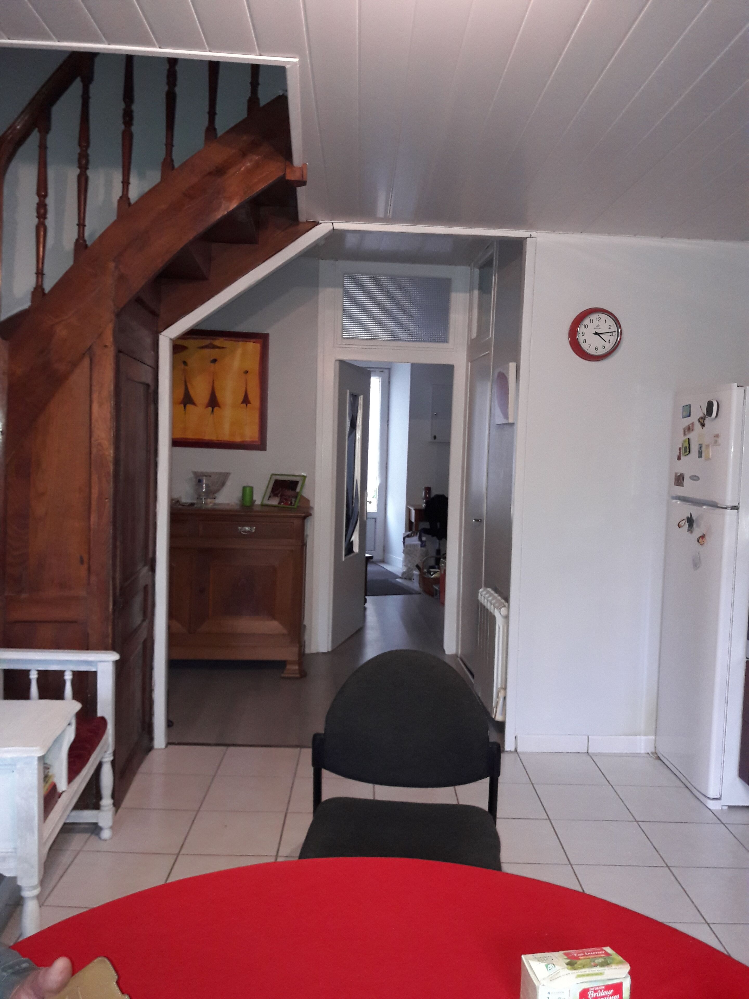 A room with a red sofa, a wooden staircase, a kitchen area, and a refrigerator.