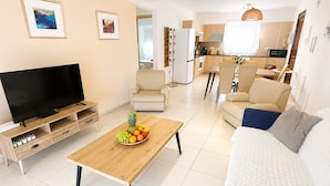 A modern living room with a flat-screen TV, a wooden coffee table with fruit, a sofa, and a dining area with chairs. - Narcissos 'Nissi Beach' Apartment D9 (Ayia Napa)