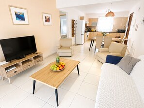 A modern living room with a flat-screen TV, a wooden coffee table with fruit, a sofa, and a dining area with chairs. - Narcissos 'Nissi Beach' Apartment D9 (Ayia Napa)