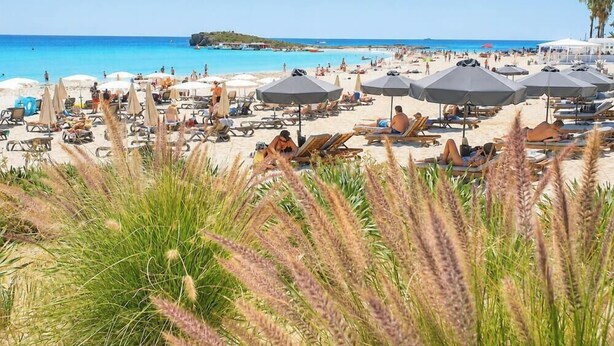 Une plage avec des chaises longues et des parasols, un ciel bleu azur et une île qui se dessine à l’horizon.