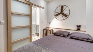 A bedroom with a bed, a wooden wardrobe with frosted glass doors, a round wall clock, a bedside table with a lamp, and a shelf with decorative items. - Le Vintage – Romantic Studio on Seine, Paris 6th (Paris)