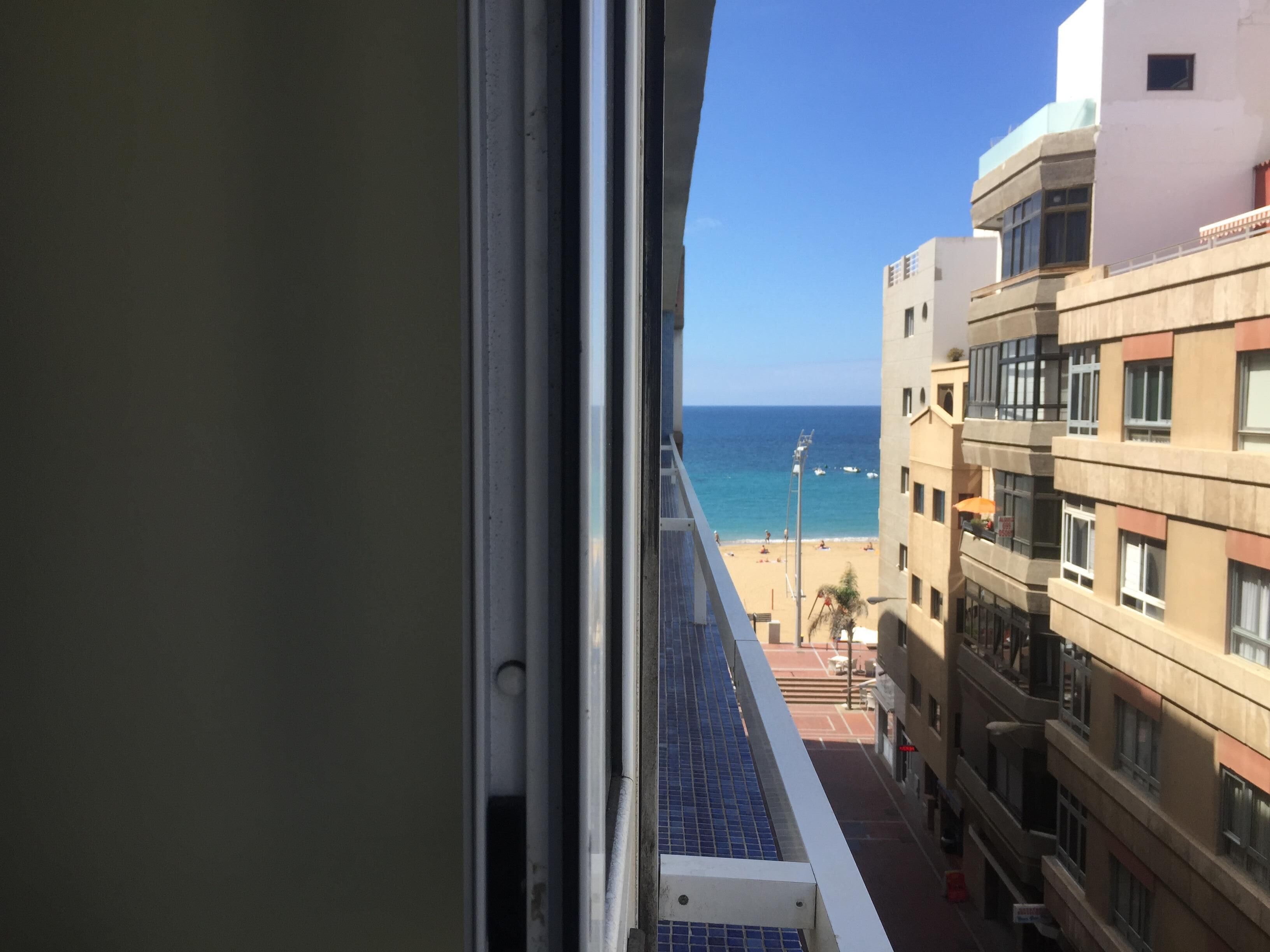 Une vue depuis une fenêtre donnant sur une plage, avec des bâtiments en arrière-plan.