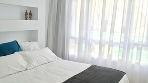 Een moderne slaapkamer met een groot bed, witte muren en uitzicht door dunne gordijnen.