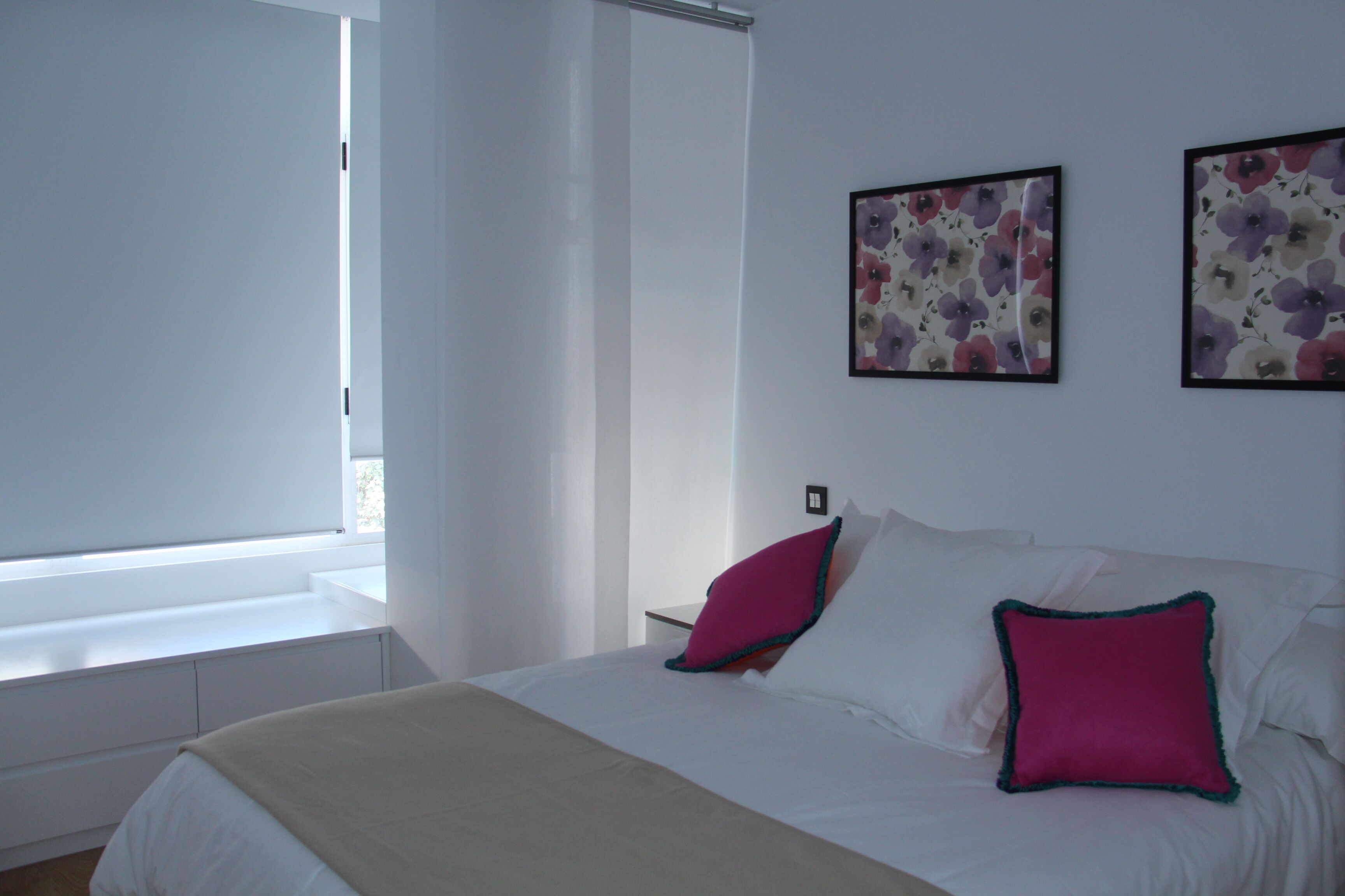 Une chambre avec une grande fenêtre, un lit avec des oreillers blancs et roses, et deux œuvres d’art florales accrochées au mur.