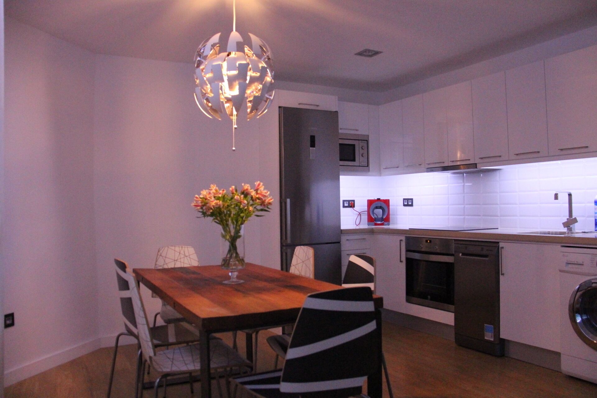 Cocina moderna con armarios blancos, un refrigerador de acero inoxidable, un microondas y una lavadora. Hay una mesa de comedor de madera con sillas y un florero con flores.