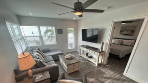 Una sala moderna con un sofá gris, una mesa de centro, un televisor y una cama en la habitación contigua.