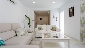 Una sala de estar moderna con un sofá color beige, una mesa de centro de vidrio y un sillón blanco. Se observa una planta en maceta y un reloj montado en la pared.