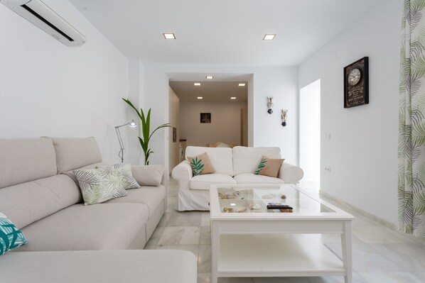 A modern living room with a beige sofa, a glass coffee table, and a white armchair. There is a potted plant and a wall-mounted clock.