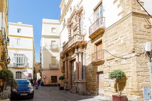 A narrow cobblestone street flanked by historic buildings with ornate balconies and a small tree in the foreground.