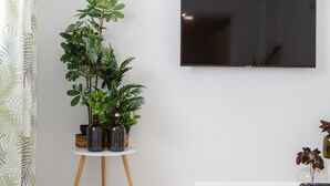 Una sala de estar moderna con un televisor de pantalla plana montado en la pared, una mesa de centro blanca con adornos y una planta en maceta sobre un pequeño pedestal.