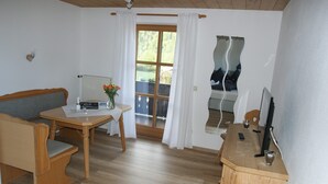 A compact room with a wooden floor, a wooden table with a chair, a sofa, a small desk with a computer, and a window with curtains.