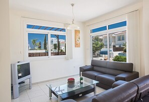A modern living room with a sofa, a glass coffee table, a TV, and large windows with blue shutters.