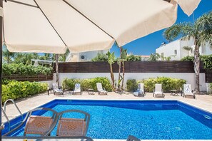 A swimming pool with lounge chairs and umbrellas, surrounded by a wooden fence and greenery.