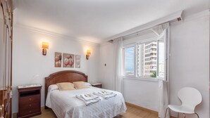 Ein Schlafzimmer mit einem hölzernen Bett, einem Nachttisch, einem Sessel und einem Fenster mit Blick auf Gebäude.