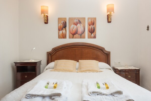Een netjes opgemaakt bed met twee kussens, een nachtkastje met een lamp, en een muur met drie schilderijen.