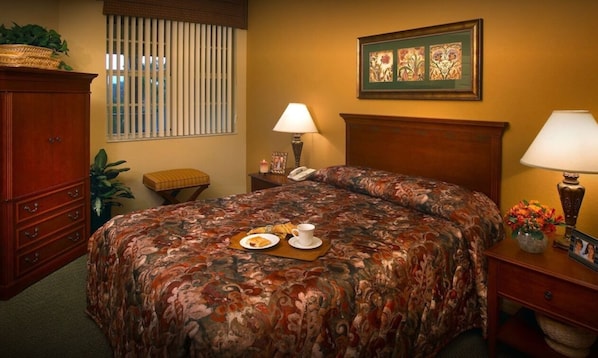 Une chambre bien rangée, avec un grand lit, deux tables de chevet avec des lampes, une petite table, une commode en bois et une fenêtre avec des stores.