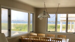 Ein Essbereich mit Tisch und Stühlen, große Fenster mit Blick auf Strand und Meer, und eine dekorative Pendelleuchte.