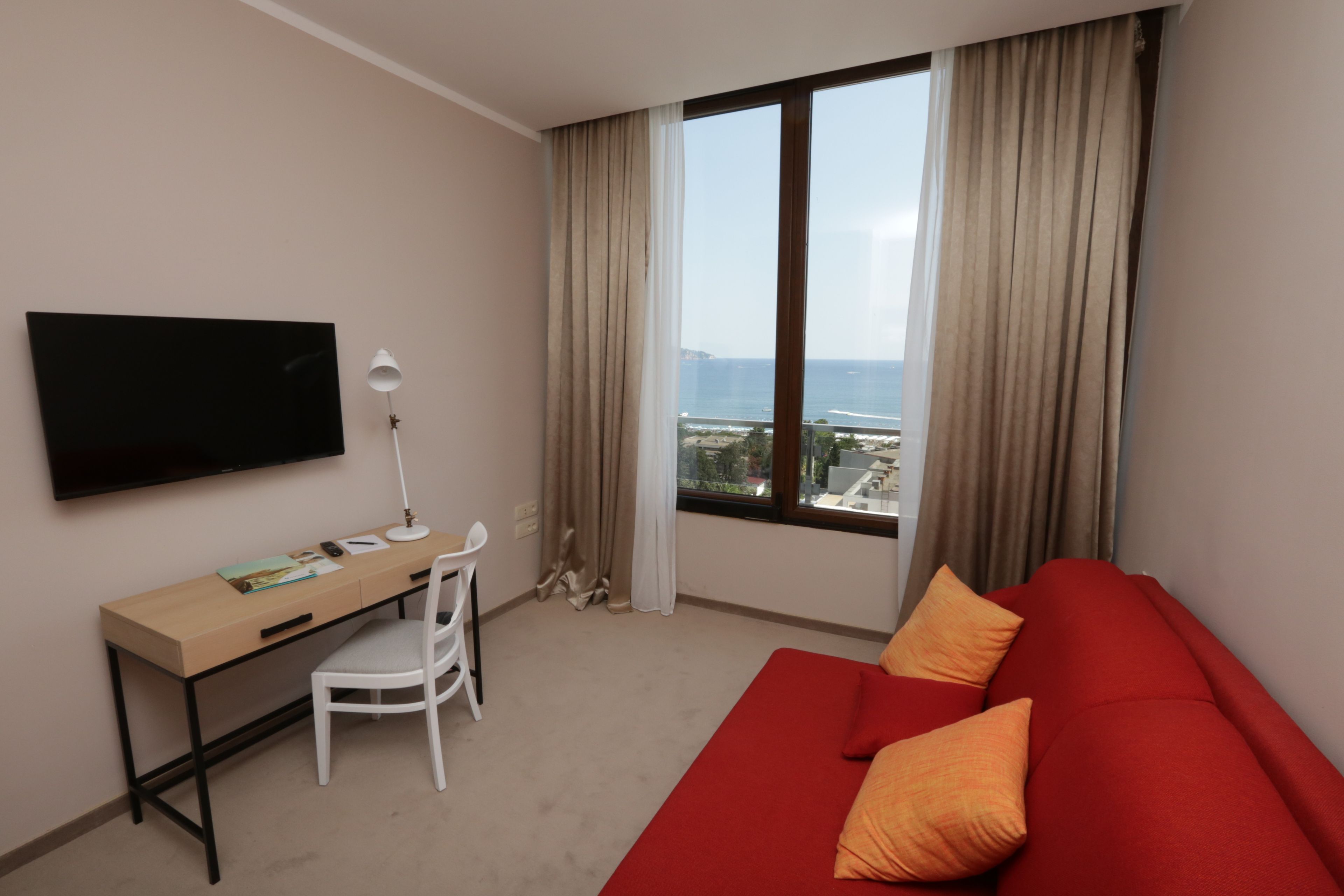 Ein Hotelzimmer mit Fernseher, Schreibtisch, Stuhl und einem roten Sofa mit orangefarbenen Kissen.