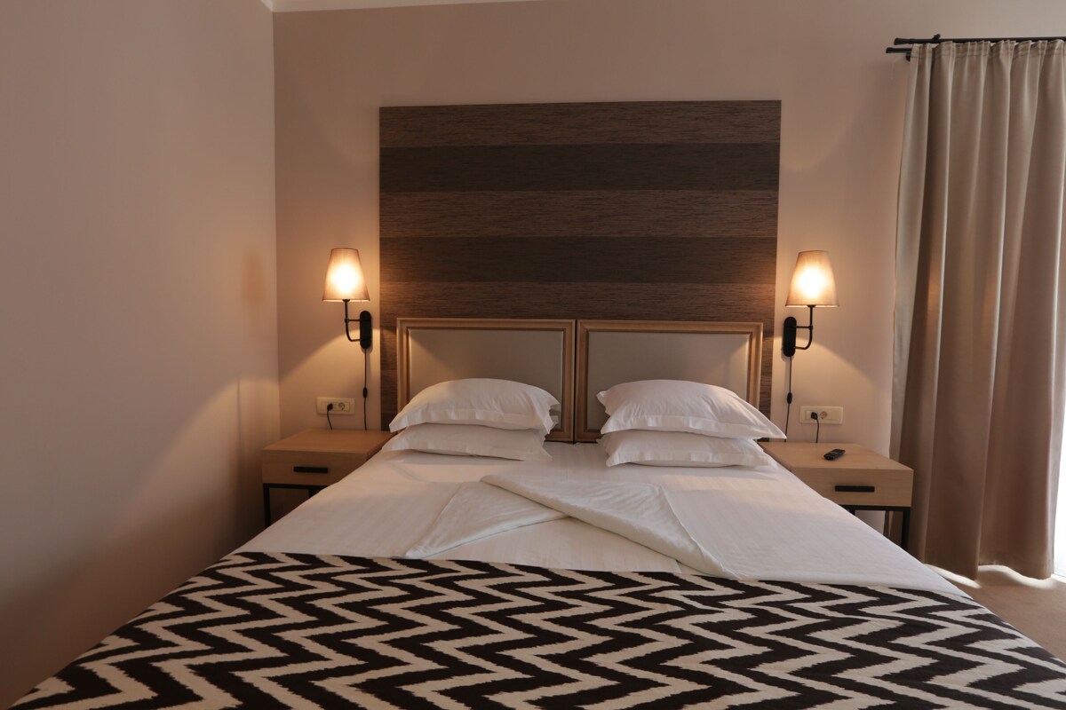A hotel room with a bed, two bedside tables, a headboard, and a window with curtains.