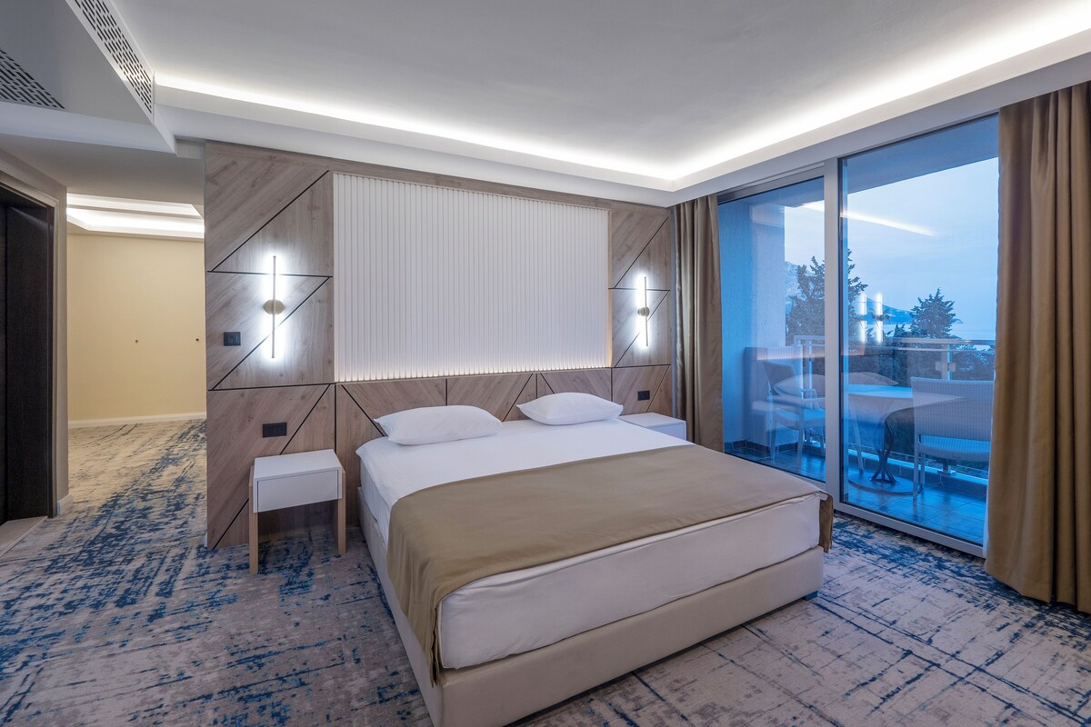A modern hotel room with a large bed, a bedside table, a nightstand, a large window with a view, and a ceiling with recessed lighting.