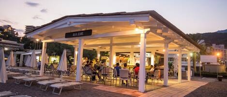 A beachside restaurant with outdoor seating, a covered area, and a sign indicating the name.